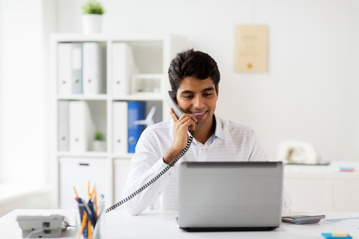 Man uses an office phone as he looks at a laptop on his desk